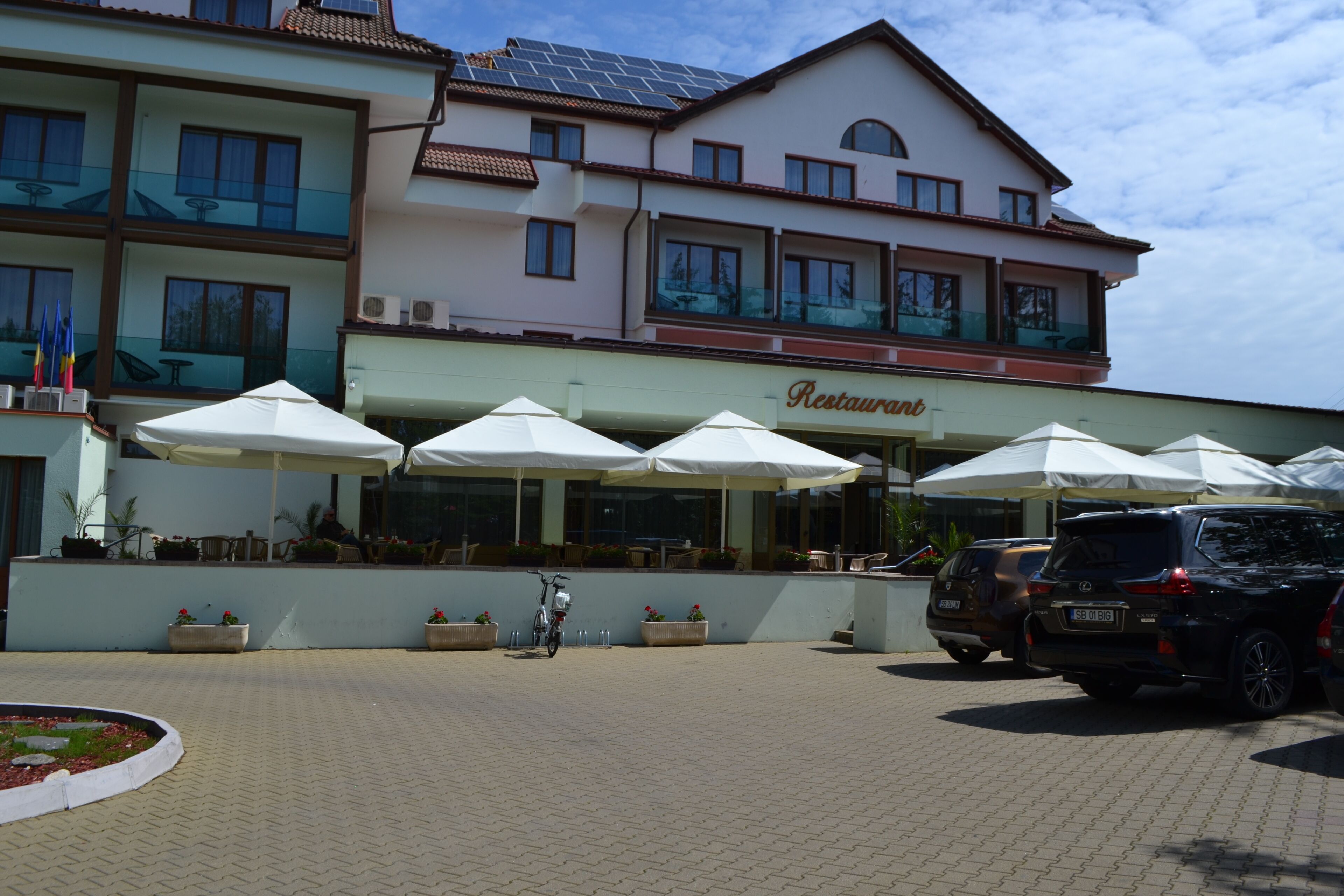 Un edificio con restaurante en la planta baja, mesas al aire libre con sombrillas y coches aparcados.