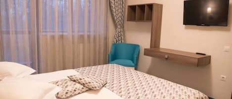 A hotel room with a bed, a blue chair, a TV mounted on the wall, and a wooden shelf.