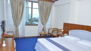 A hotel room with a large bed, a wooden nightstand, a desk with a chair, a small table with a lamp, and a view of the city through a window with curtains. - Panorama Hotel (Yangon)