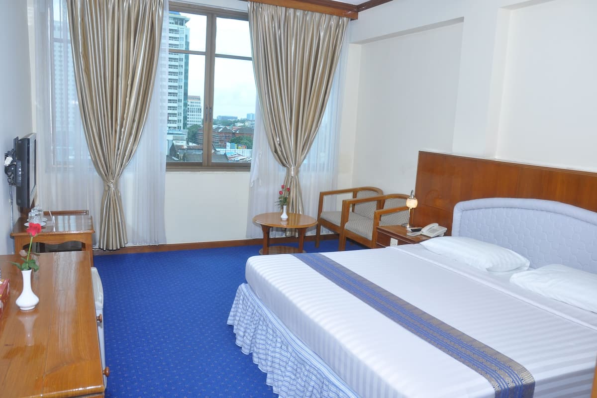 A hotel room with a large bed, a wooden nightstand, a desk with a chair, a small table with a lamp, and a view of the city through a window with curtains.