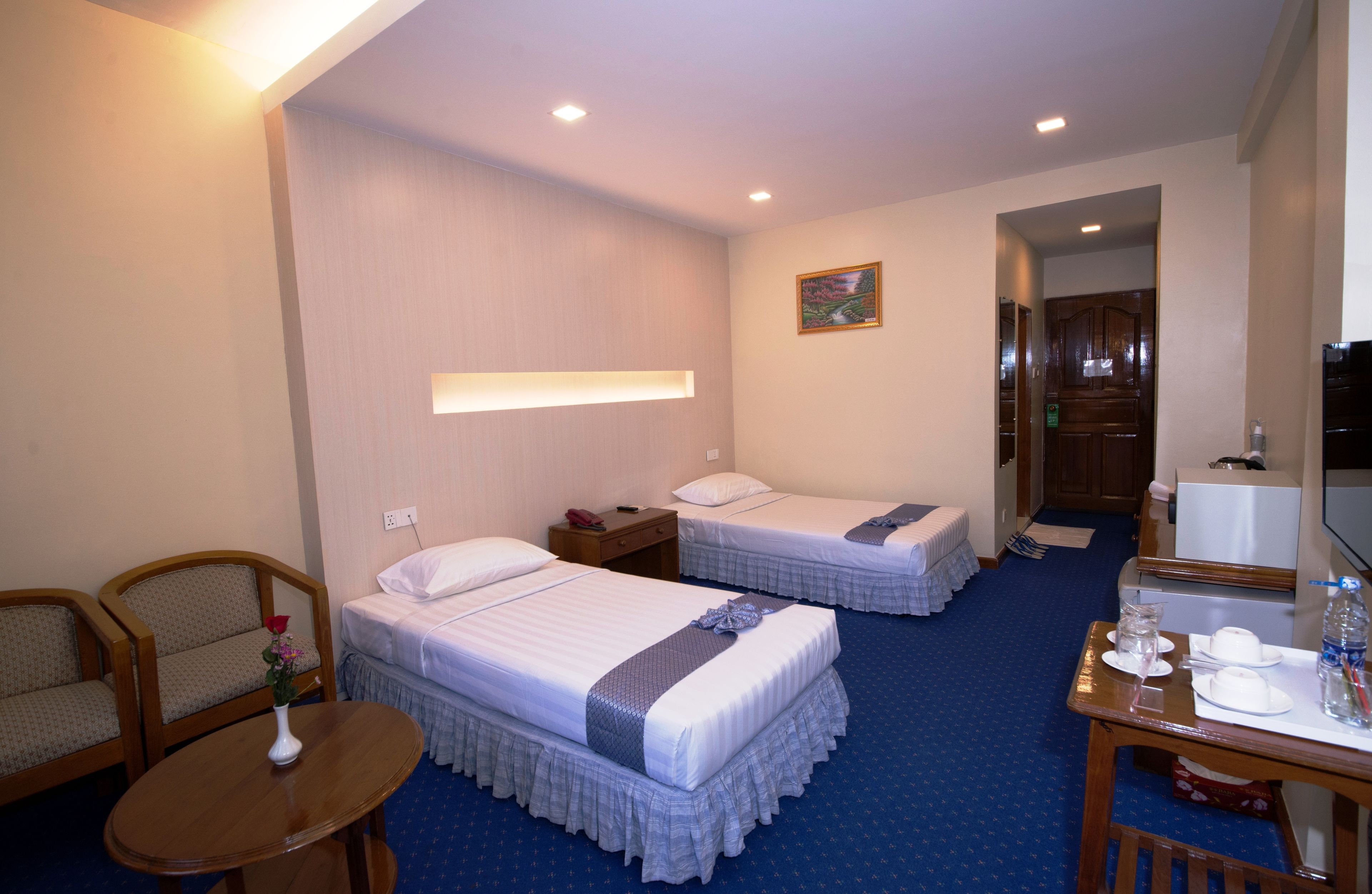 Habitación de hotel con dos camas, un escritorio con silla, una mesita pequeña, un florero con flores, un cuadro en la pared y una puerta que da al baño.
