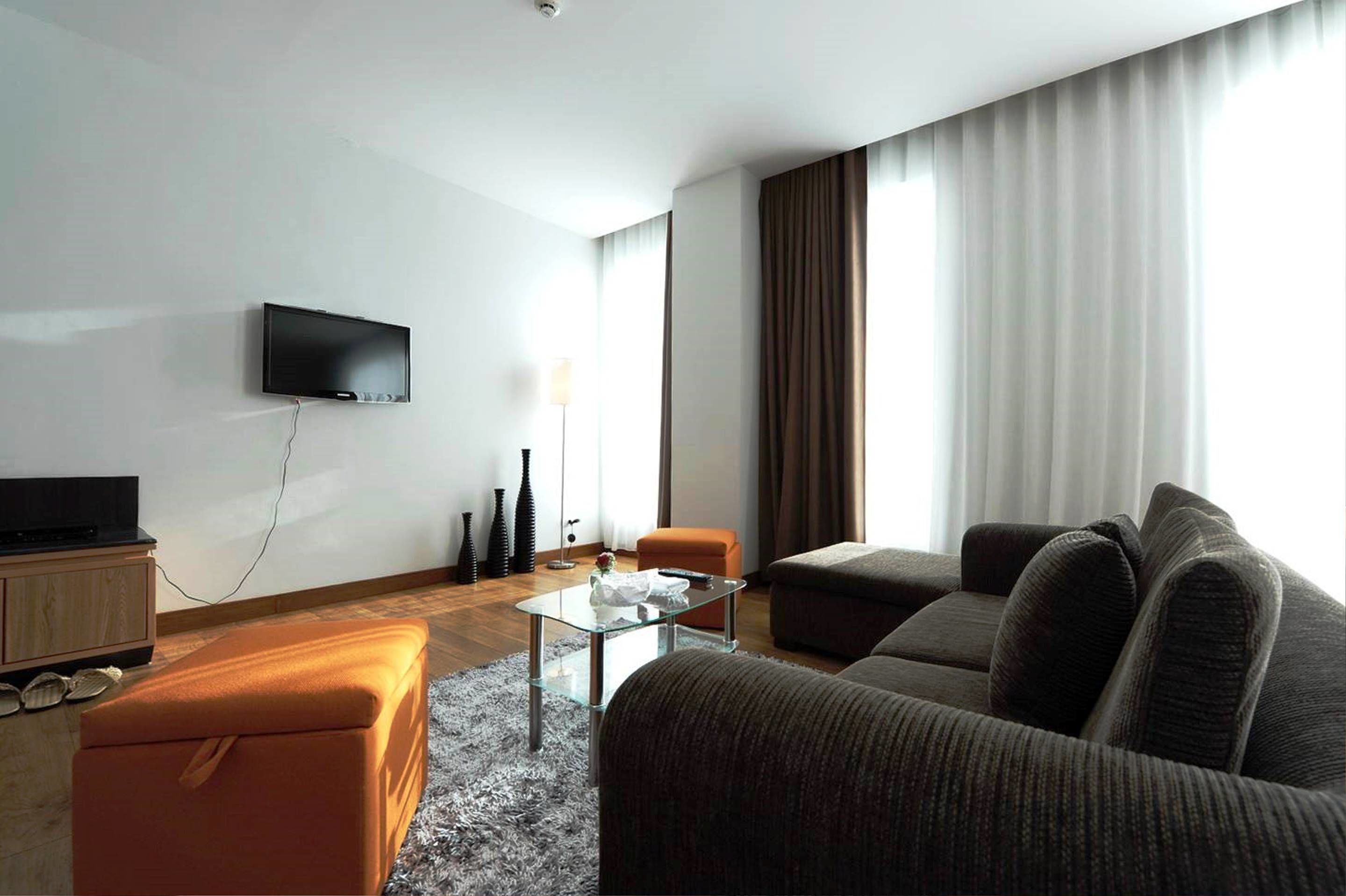 A modern living room with a flat-screen TV mounted on the wall, a glass coffee table, a sofa, an ottoman, and a lamp.
