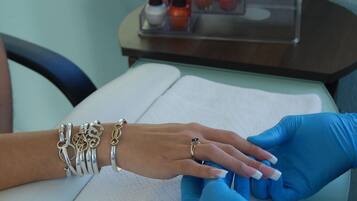 A close-up of a person's hand with manicured nails, wearing a silver bracelet and a ring, being attended to by a person in blue gloves.