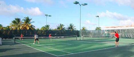 A tennis court with a net, palm trees, and street lamps.