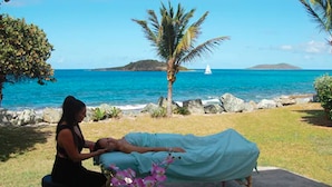 A person lying on a massage table outdoors with a view of the ocean and a sailboat in the distance.