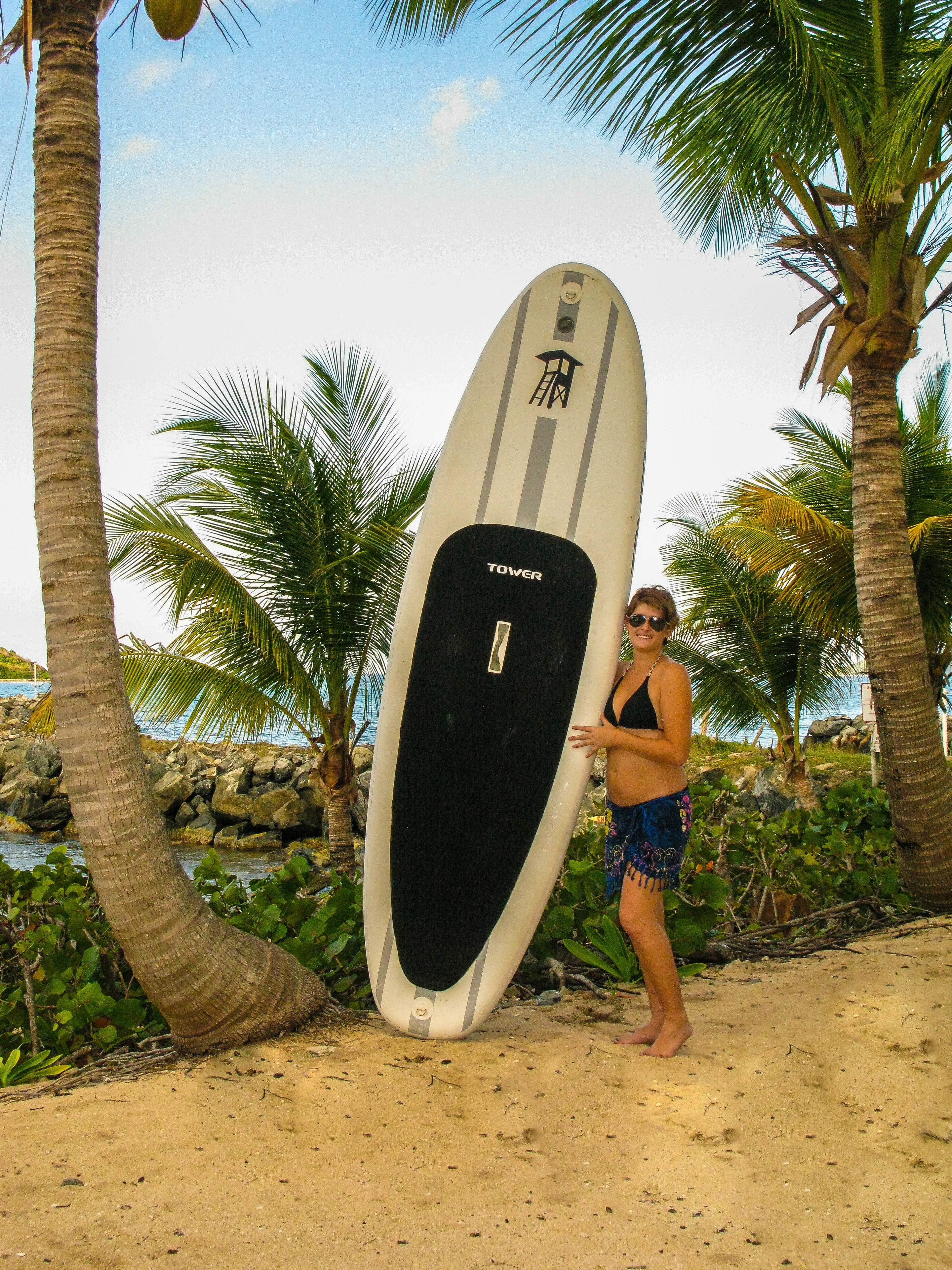 A person holding a surfboard on a sandy beach with palm trees and a rocky shoreline in the background.