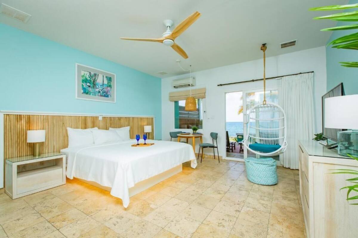 Habitación de hotel con cama, ventilador de techo, sillón colgante y vista al océano.