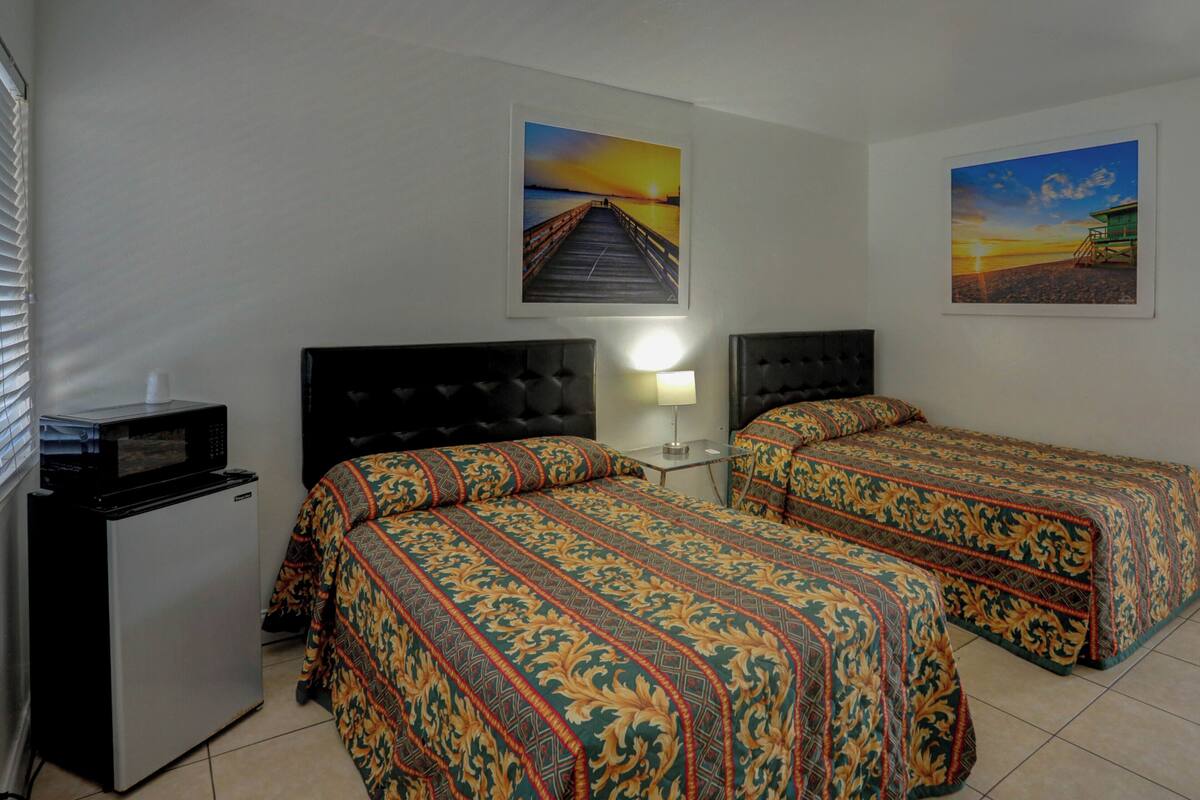 A hotel room with two beds, a small refrigerator, a microwave, and two wall-mounted paintings.