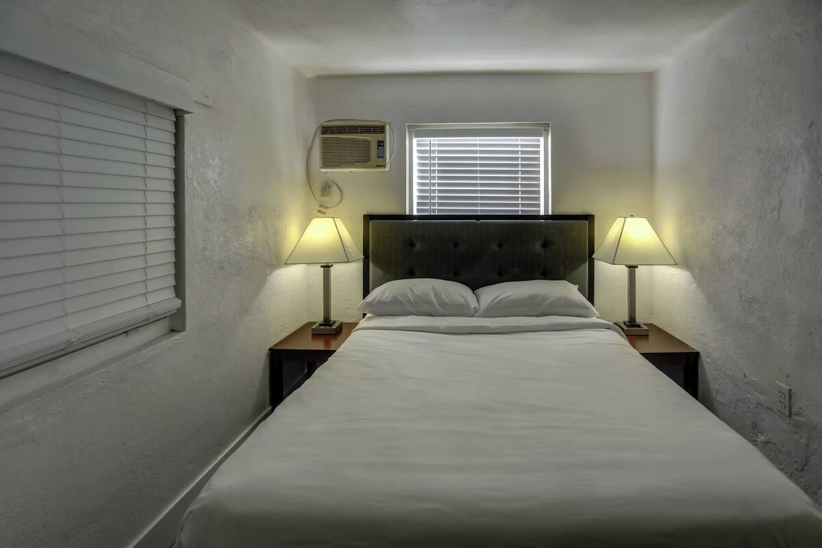 A neatly made bed with two bedside lamps, a window with blinds, and an air conditioning unit on the wall.