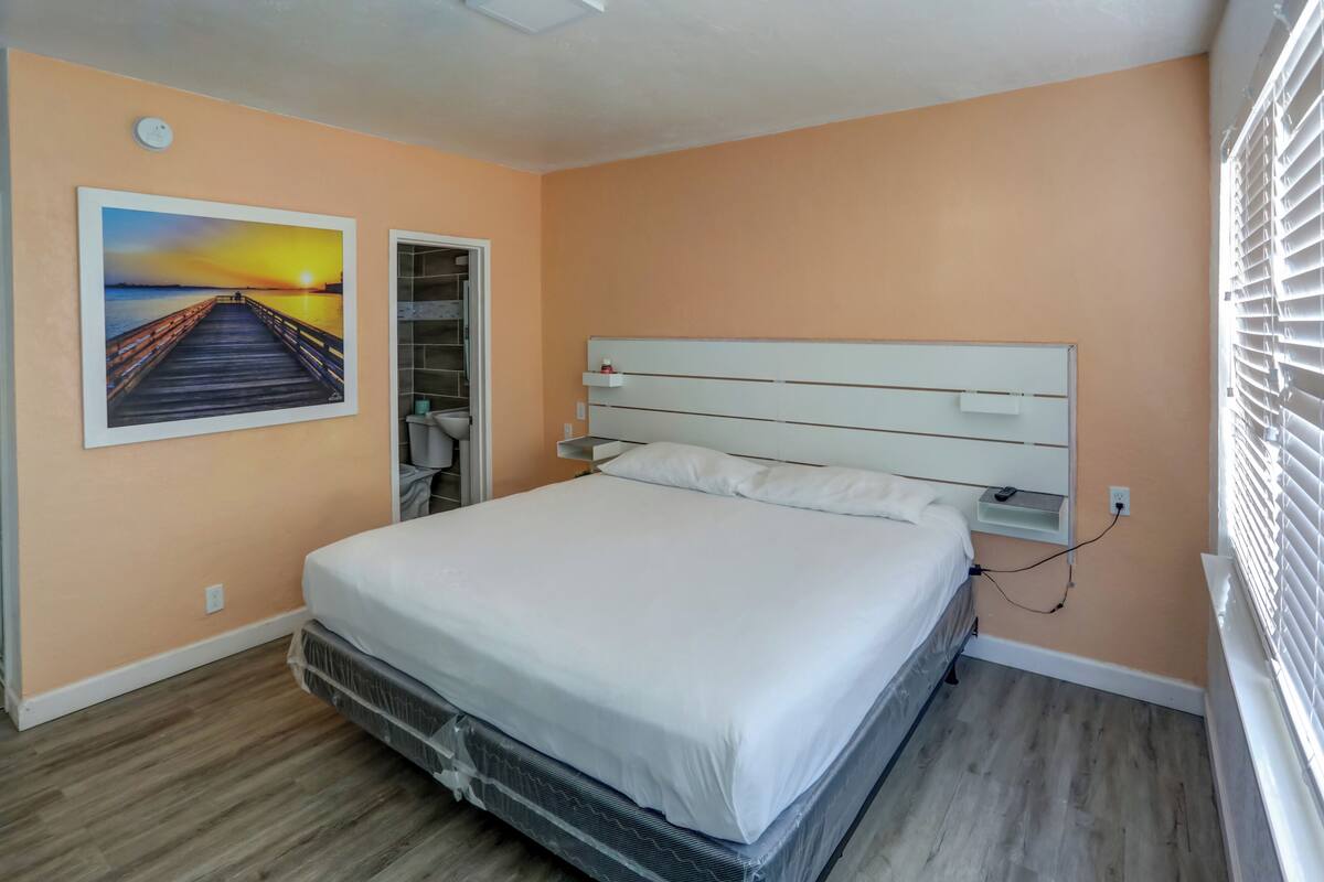 A bedroom with a bed, a nightstand, a wall-mounted shelf, a painting, and a window with blinds.