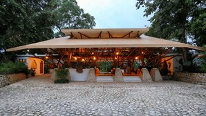 A stone-paved courtyard with a wooden pavilion supported by a metal framework, surrounded by trees and a stone wall. - Hotel Villa Maya (Flores)