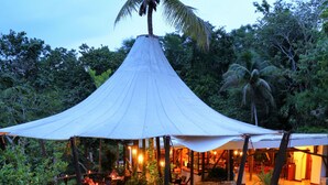 A covered outdoor dining area with tables and chairs, surrounded by greenery and a palm tree. - Hotel Villa Maya (Flores)
