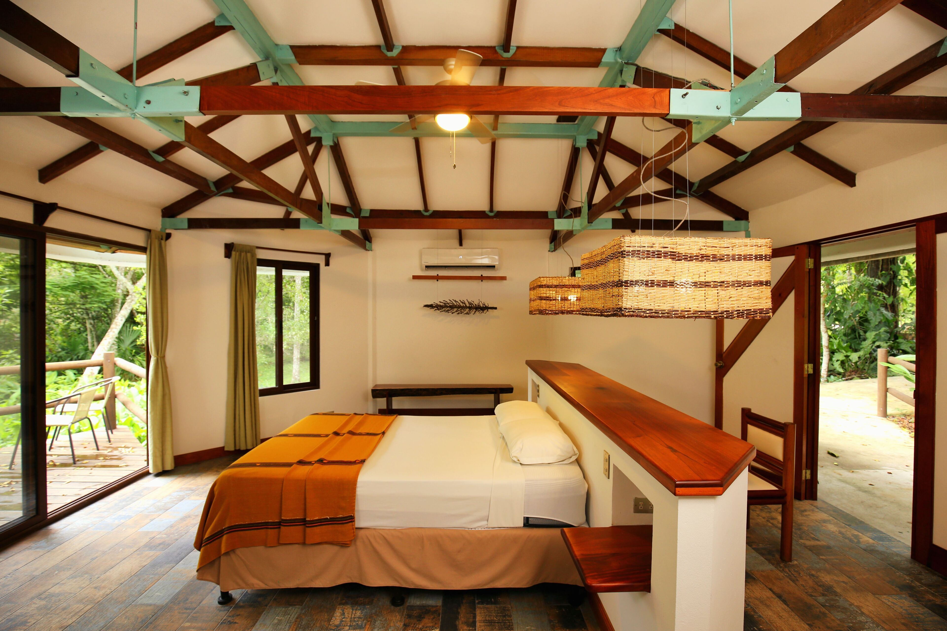 A bedroom with a bed, wooden ceiling beams, an air conditioning unit, and a view through a glass door.