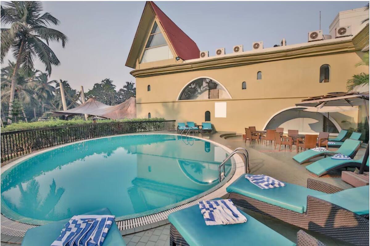 Una piscina con sillas de descanso y sombrillas, un edificio de techo triangular y palmeras al fondo.