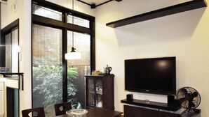 A modern living room with a dark wooden dining table, cushioned benches, a flat-screen TV, and a large window with a view of greenery. - Kyoto Guesthouse Roujiya (Kyoto)