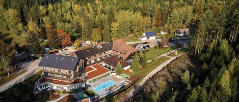 A large building with multiple sections, a swimming pool, and a parking area surrounded by dense forest.