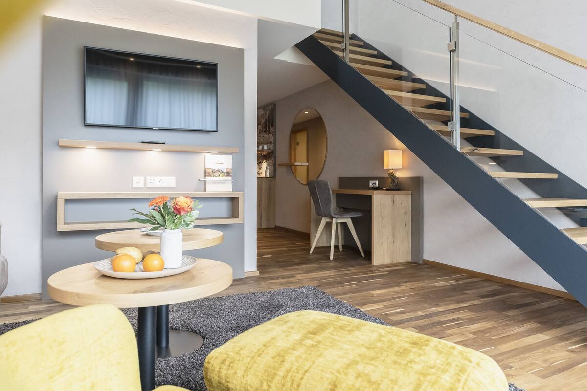 A modern living room with a wooden floor, a round coffee table with oranges and a vase, a TV mounted on the wall, and a staircase with a glass railing.