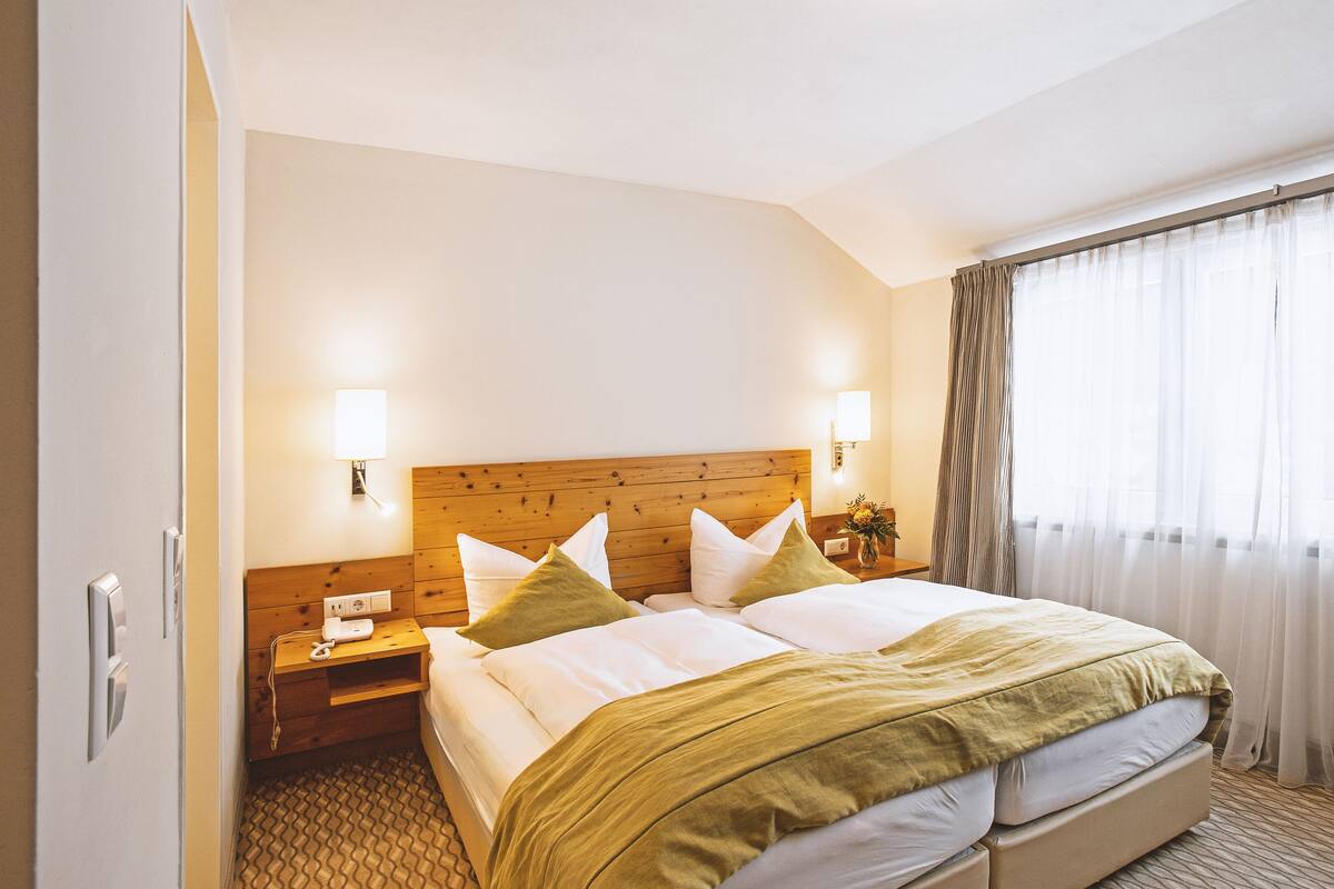 A hotel room with a wooden headboard, two bedside tables with lamps, a large bed with white and green bedding, and a window with sheer curtains.