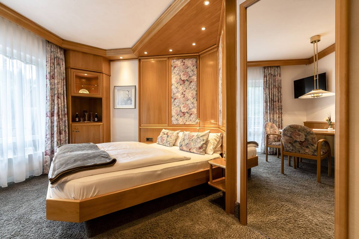 A hotel room with a large bed, wooden headboard, a desk with a chair, a television, and a floral-patterned wall panel.