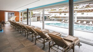 A row of wicker lounge chairs with cushions facing a swimming pool, with a view of a snowy outdoor area. - Hotel Restaurant Waldsägmühle (Pfalzgrafenweiler)