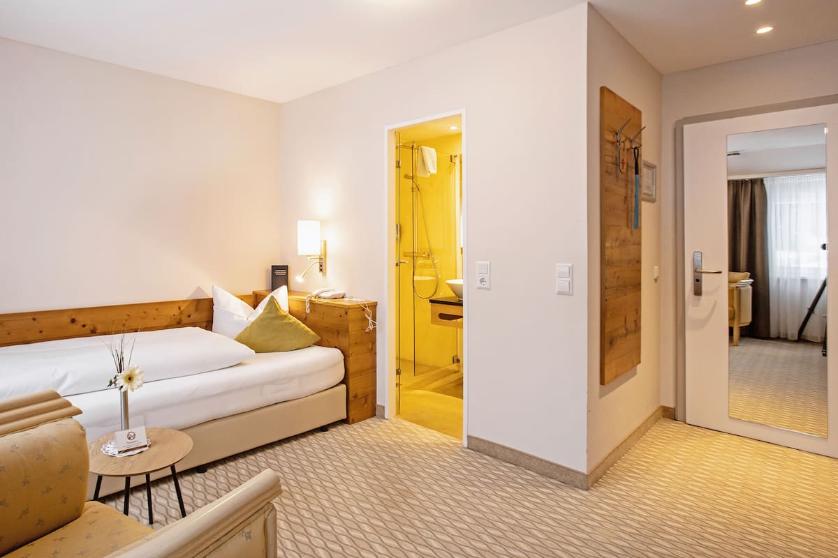 A hotel room with a bed, a bedside table, a chair, a small table, a mirror, and a bathroom door.