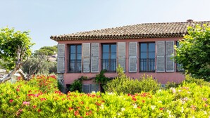 Un edificio rosa con persianas y techo de tejas, rodeado de abundante vegetación y plantas con flores.