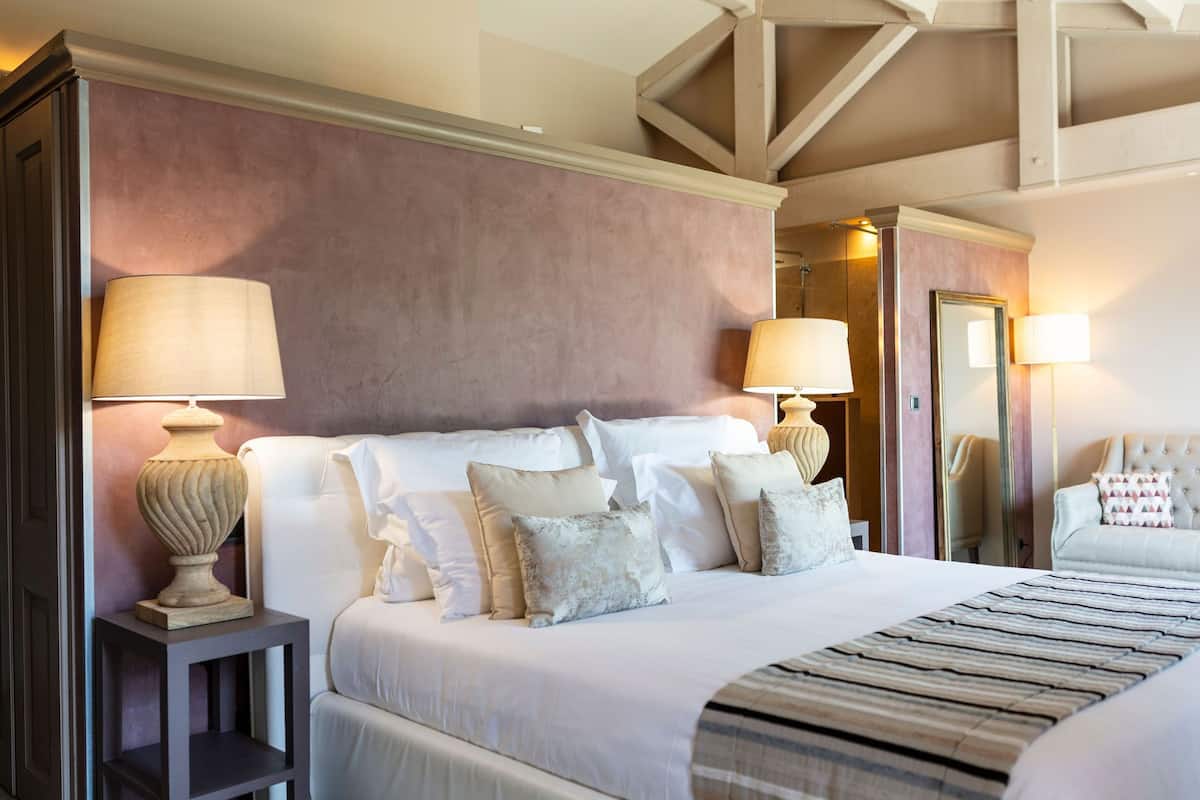 A neatly made bed with white linens and multiple pillows, a bedside lamp, and a small table with a lamp.