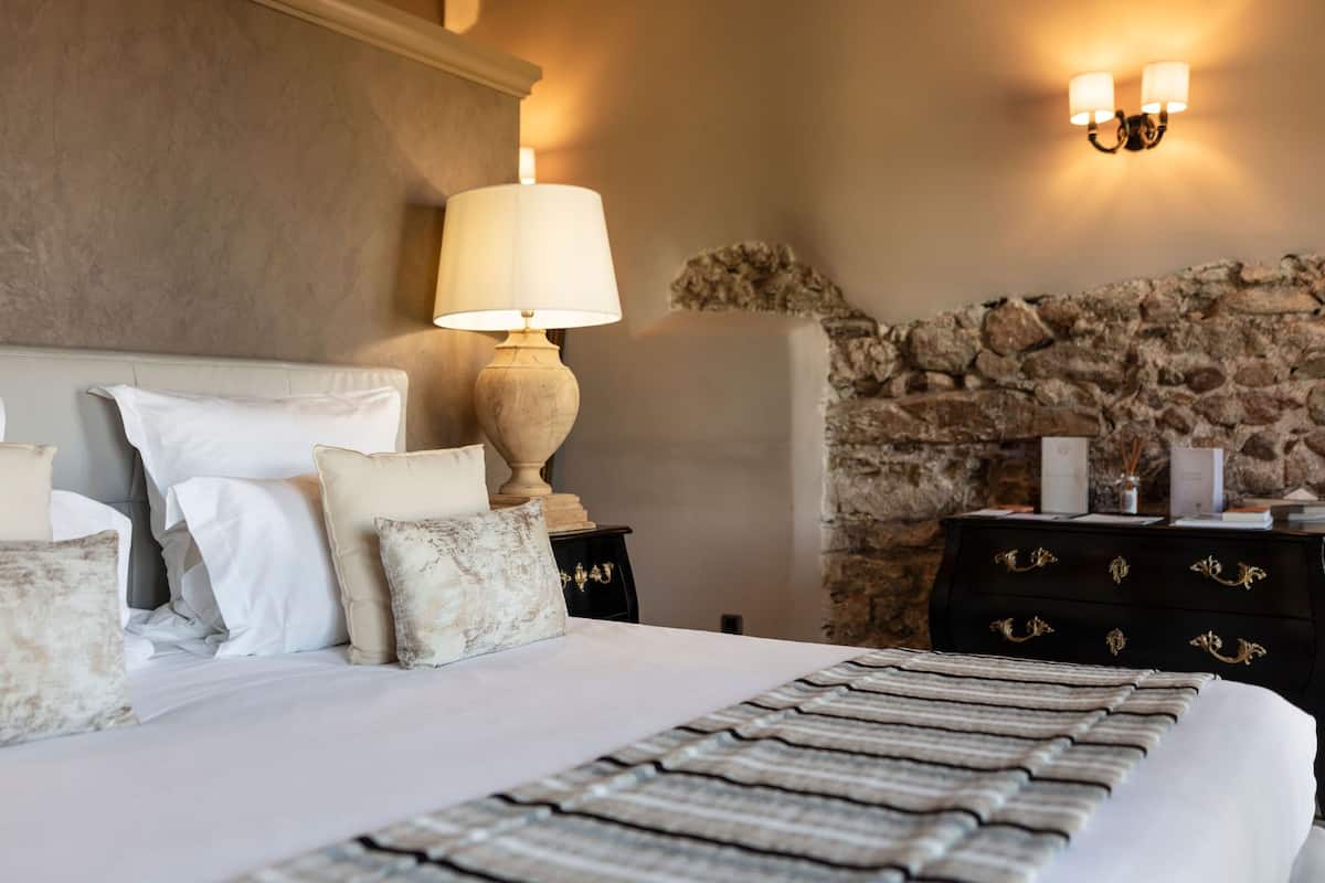 A neatly made bed with white linens and decorative pillows, a stone wall, and a nightstand with a lamp and decorative items.