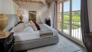 Un dormitorio con una cama grande, una mesita de noche, una lámpara, un espejo y vistas a un viñedo a través de amplios ventanales.