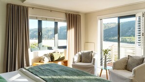 A hotel room with a bed, two chairs, a small table, and a view of a mountainous landscape through large windows. - Christiana Lodge (Plettenberg Bay)