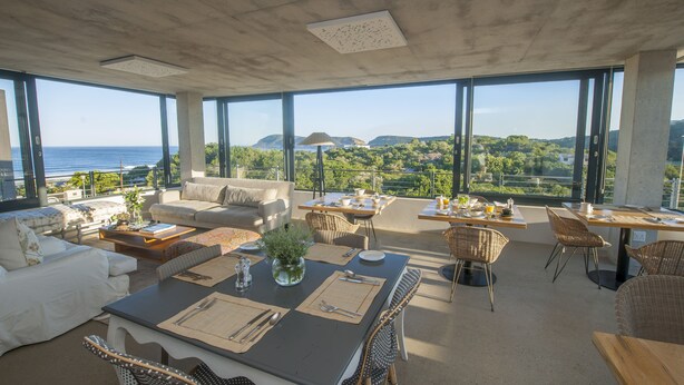 A spacious living area with a large dining table, comfortable seating, and a view of the ocean. - Christiana Lodge (Plettenberg Bay)