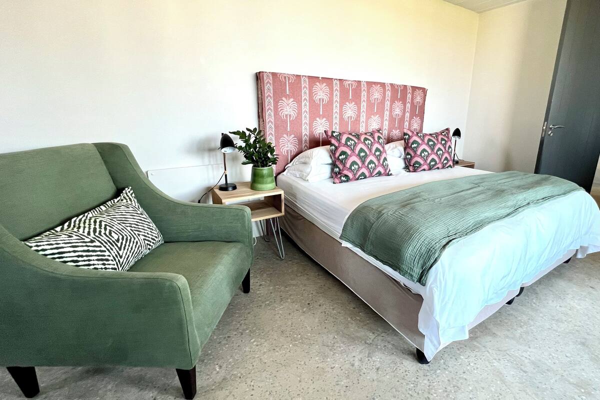 A bedroom with a green armchair, a bed with patterned pillows, a bedside table with a lamp, and a green plant.