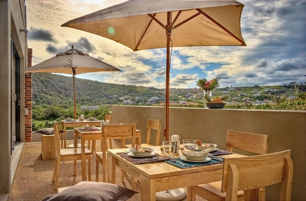 Un espace de restauration en plein air, doté de mobilier en bois et de grands parasols, offrant une vue panoramique sur une colline pittoresque.