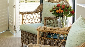 A wicker chair with a cushion, a vase with flowers, and a view of greenery through a glass door. - Christiana Lodge (Plettenberg Bay)