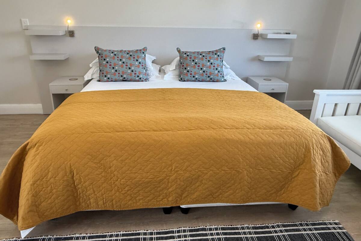 A neatly made bed with a mustard-colored quilt, two patterned pillows, and a white headboard. There are two bedside tables with lamps, and a white chair to the right.
