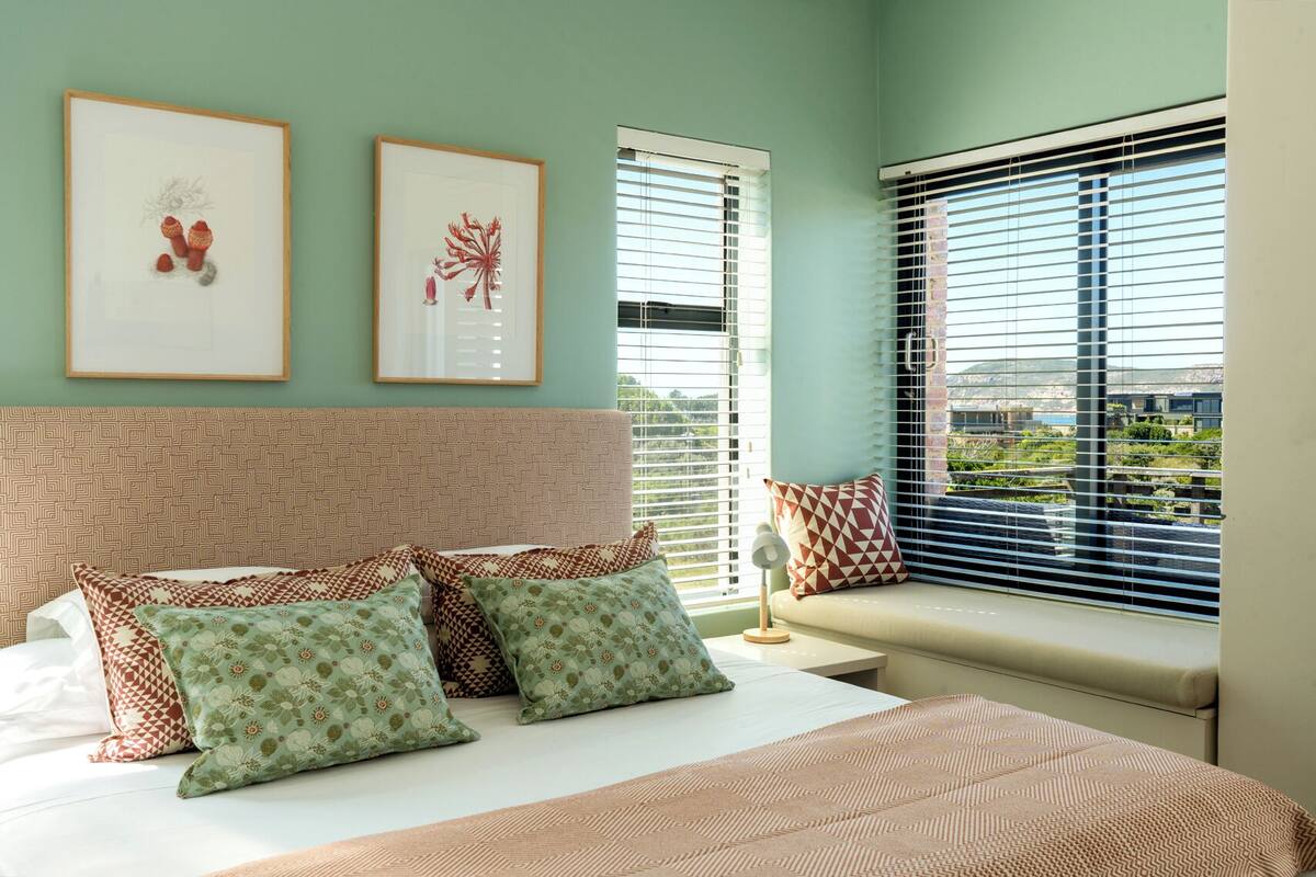 A bedroom with a bed, pillows, a headboard, two framed pictures, a window with blinds, and a view of greenery outside.