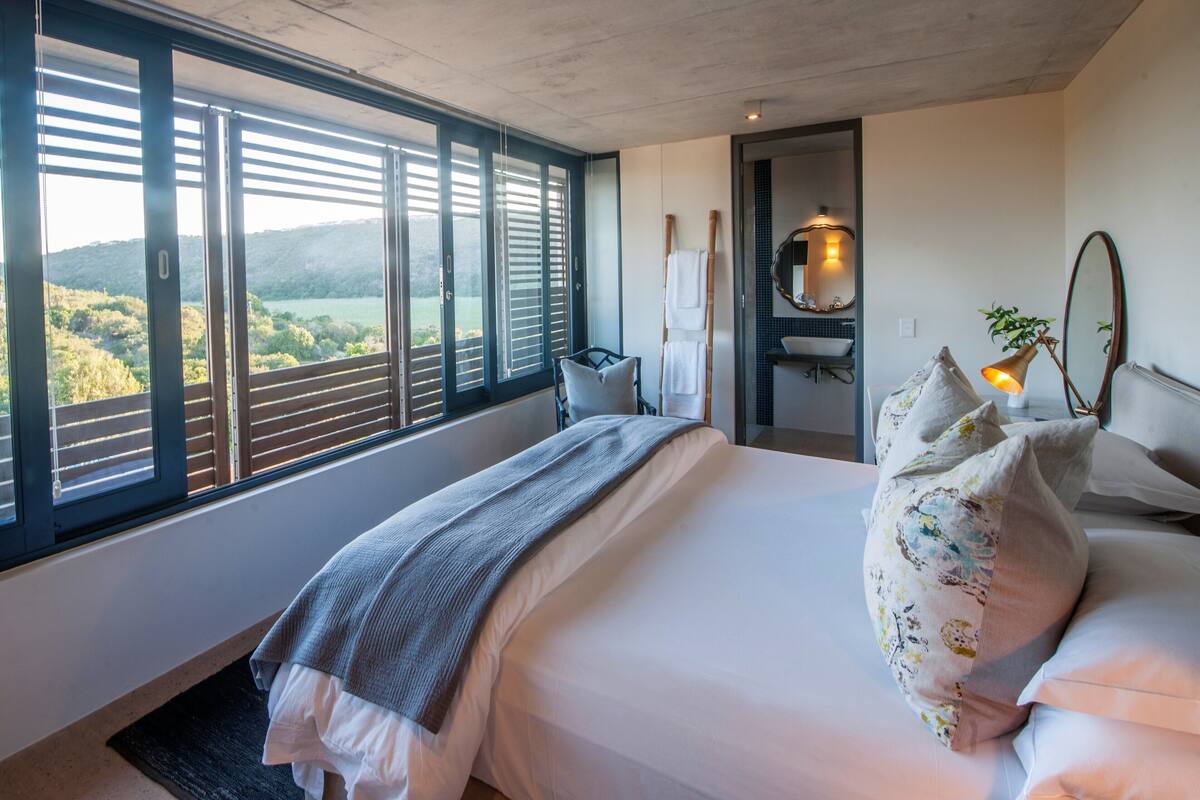 A bedroom with a large window, a bed with white linens, a bedside table with a lamp, and a view of a green landscape.