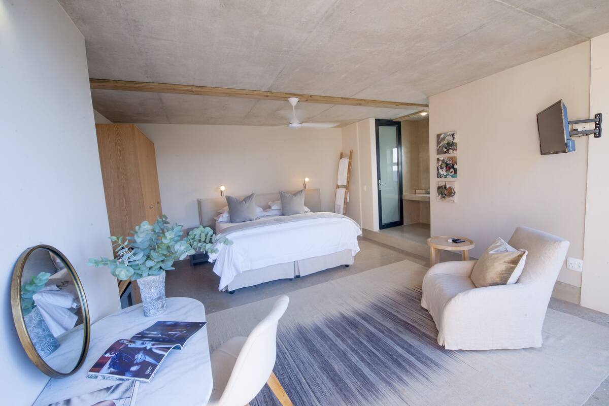 A modern bedroom with a large bed, bedside tables, a chair, a mirror, and a small table with a plant.