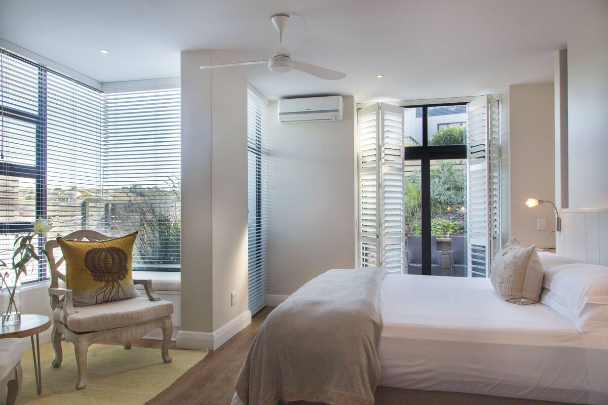 A bedroom with a bed, a chair, a window with blinds, an air conditioning unit, and a ceiling fan.