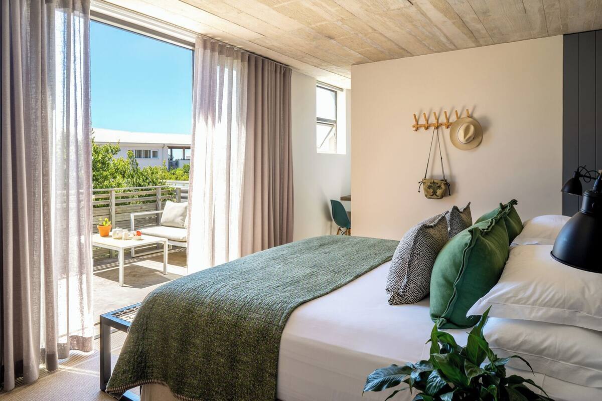 A bedroom with a bed, pillows, a bedside lamp, a hanging hat rack, and a view of the outdoors.