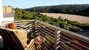A balcony with a wicker chair and a table, overlooking a river and a hilly landscape. - Christiana Lodge (Plettenberg Bay)