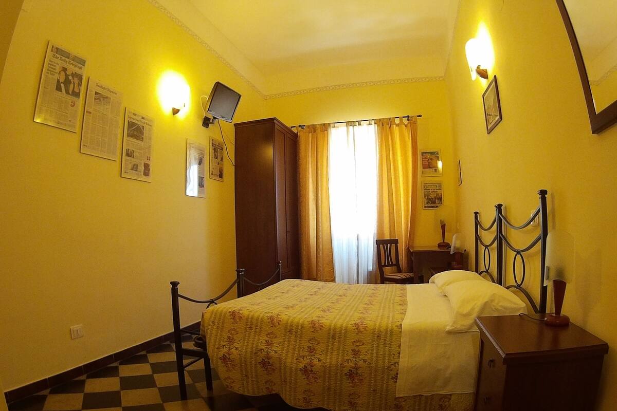 A hotel room with a checkered floor, a bed with a floral bedspread, a nightstand, a desk with a chair, and a wall-mounted TV.