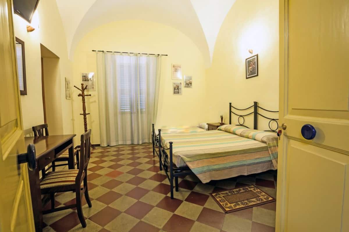 A room with a checkered floor, a bed with a striped bedspread, a wooden chair, a small table, and a door with a blue handle.
