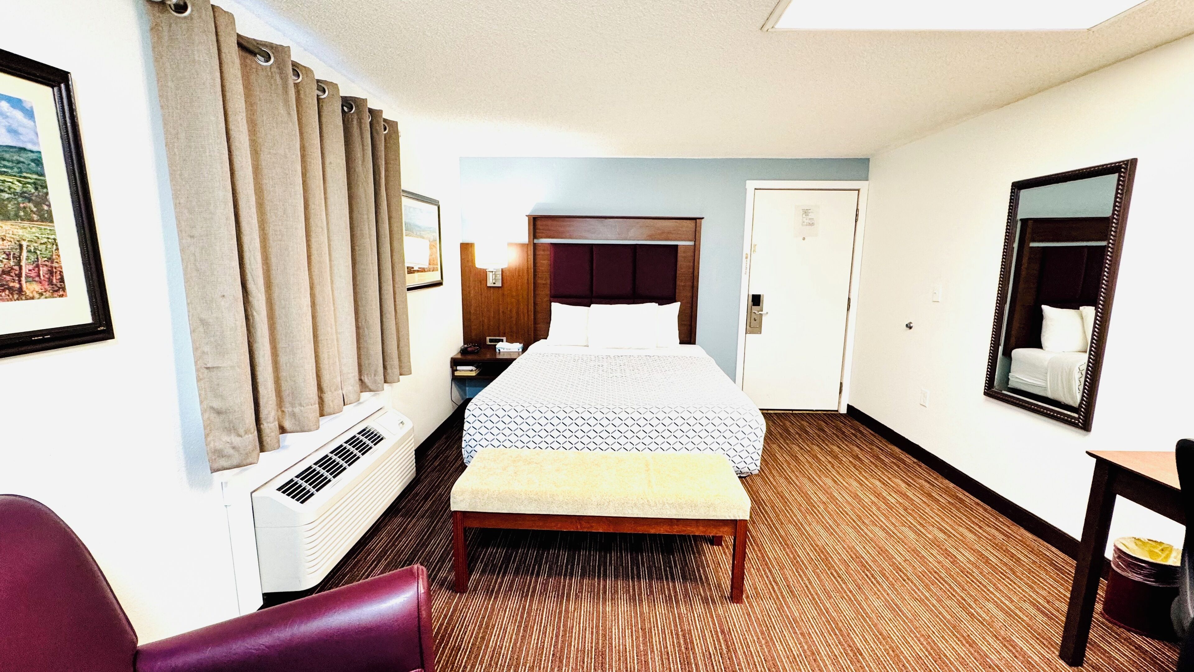 A hotel room with a bed, a bench, a desk, a chair, a mirror, a picture frame, and a window with curtains.