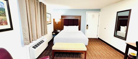 A hotel room with a bed, a bench, a desk, a chair, a mirror, a picture frame, and a window with curtains.
