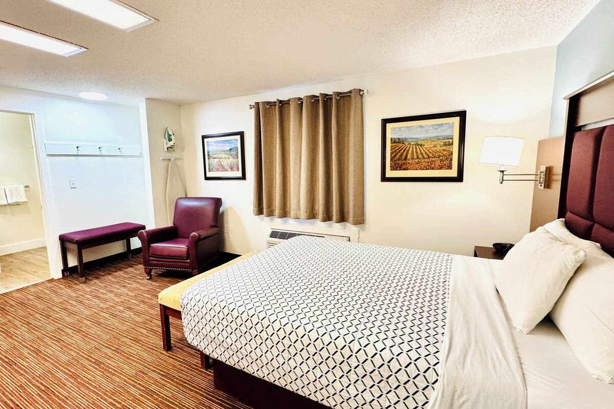 A hotel room with a bed, a chair, a desk, a window with curtains, and a painting on the wall.
