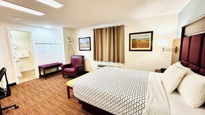 A hotel room with a bed, a chair, a desk, a window with curtains, and a painting on the wall.