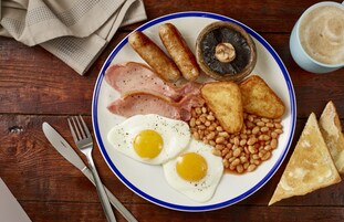 A plate with eggs, sausages, mushrooms, bacon, beans, toast, and a cup of coffee on a wooden table.
