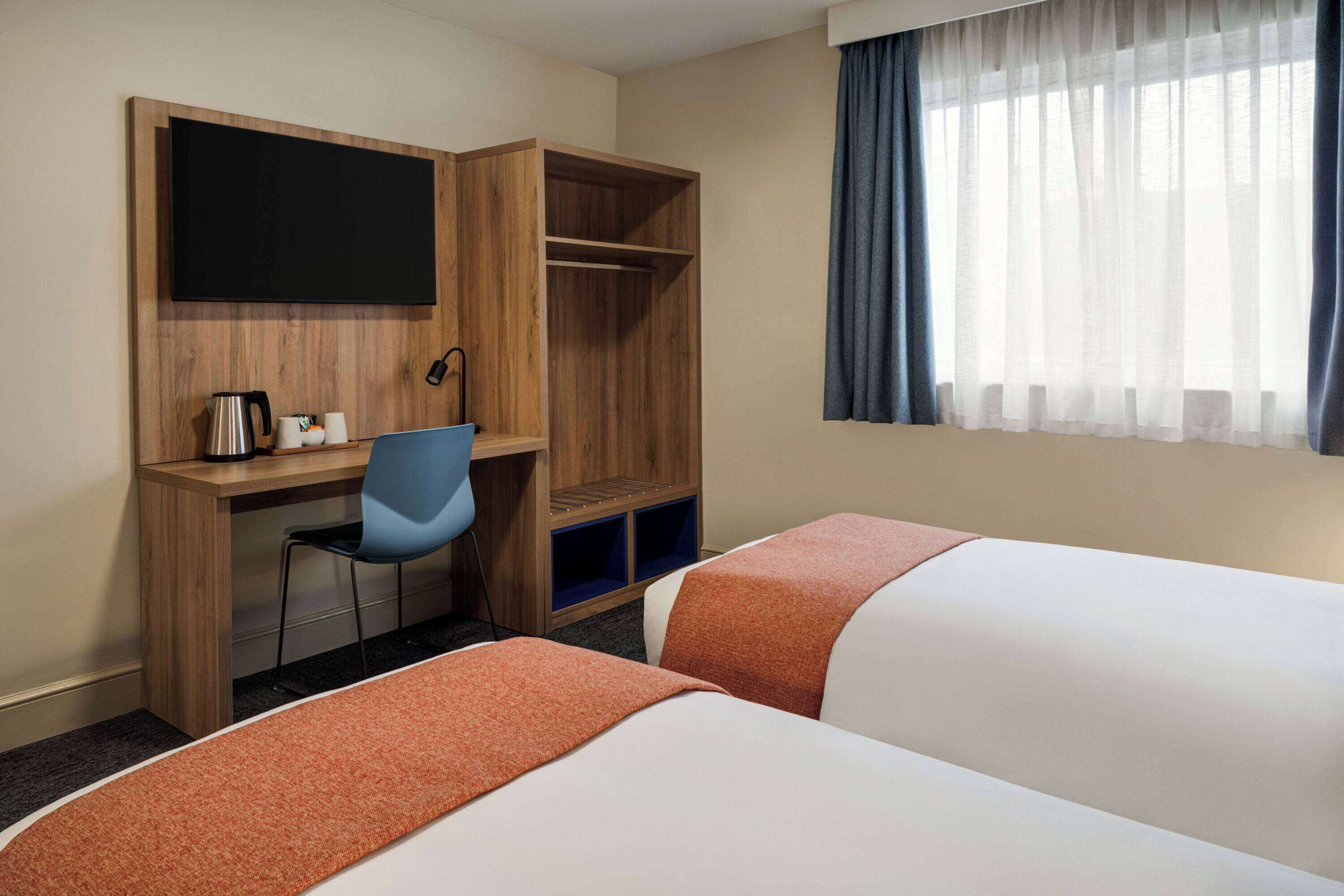 A hotel room with two beds, a desk with a chair, a television, and a window with curtains.
