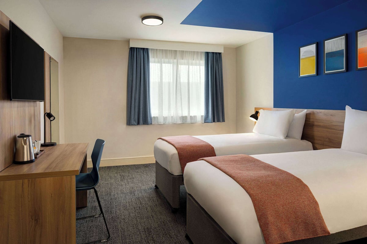 A hotel room with two beds, a desk, a chair, a TV, and a window with curtains.